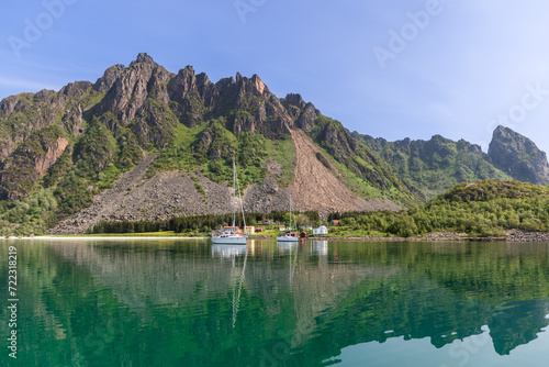 A tranquil summer scene in the Lofoten Islands, where sailboats and traditional Rorbu cabins are mirrored in the still waters under a rugged mountainous backdrop. Norway