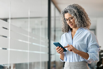 Happy middle aged business woman holding mobile cell phone using cellphone in office. Smiling mature old professional lady business owner entrepreneur using smartphone standing in hallway.