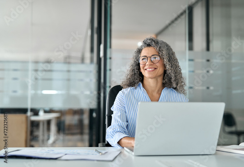 Happy middle aged professional business woman working in office with laptop looking away. Smiling older lady executive corporate leader at workplace thinking of new business ideas. Copy space.