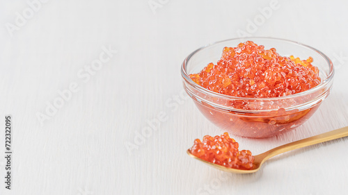 Salty natural salmon red caviar or organic fish roe served in transparent glass bowl on white wooden background with copy space with spoon of gold colour used as spread or ingredient for breakfast