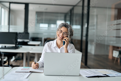Happy middle aged business woman talking on phone using laptop in office. Smiling mature older female bank manager, senior professional lady making business call on telephone working sitting at desk.