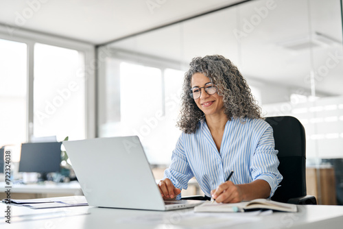 Busy mature business woman working in office using laptop writing. Smiling middle aged professional executive looking at computer, elearning, watching webinar having hybrid meeting sitting at desk.