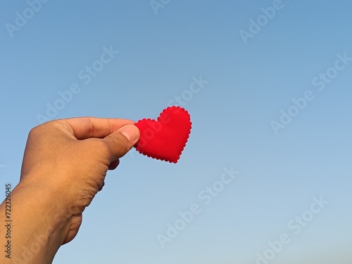 Red heart with hand under blue sky and sun light background, love and valentines day concept.