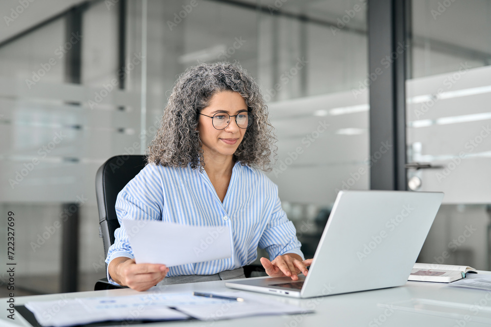 Busy mature business woman working in office looking at laptop holding document. Older professional hr manager, lawyer or notary checking financial tax report account in office. Business consultancy. Busy mature business woman working in office looking at laptop holding document. Older professional hr manager, lawyer or notary checking financial tax report account in office. Business consultancy.