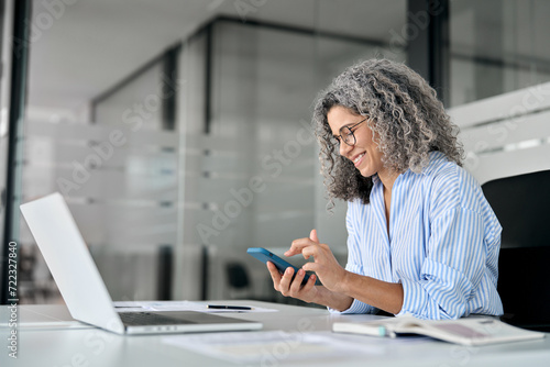 Happy older mature business woman holding mobile phone using cellphone in corporate office. Smiling middle aged professional lady executive using smartphone working on cellular sitting at desk. photo