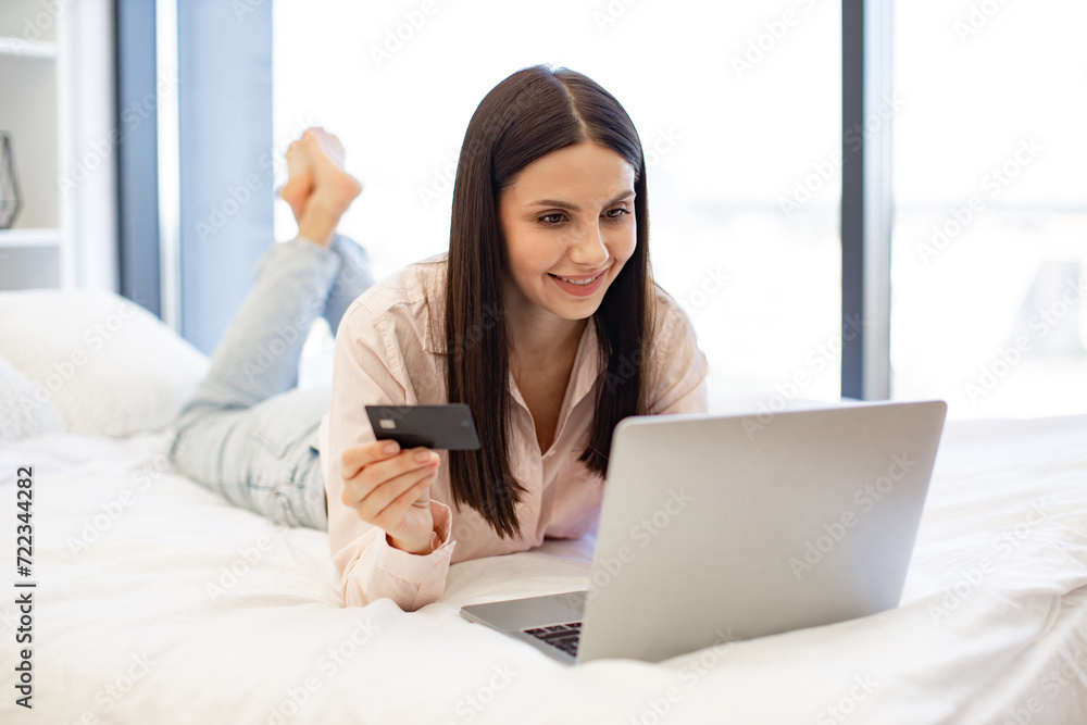 Smiling young woman making online order using modern wireless laptop. Relaxed lady lying on comfy bed and enjoying purchase of goods on Internet using bank card at home on weekend. Smiling young woman making online order using modern wireless laptop. Relaxed lady lying on comfy bed and enjoying purchase of goods on Internet using bank card at home on weekend.