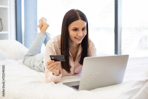 Smiling young woman making online order using modern wireless laptop. Relaxed lady lying on comfy bed and enjoying purchase of goods on Internet using bank card at home on weekend.