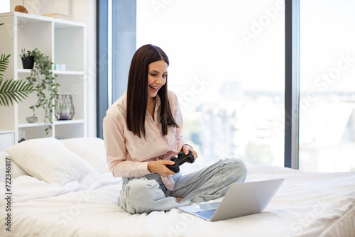 Happy young woman holding wireless joystick while playing video games on modern laptop. Happy lady sitting on comfy bed and enjoying favorite computer game on weekend.