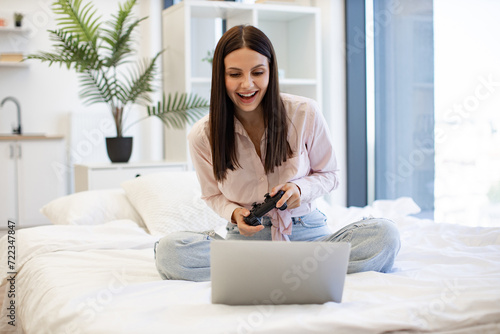 Caucasian brunette using modern devices enjoying favorite video game on weekend. Beautiful young woman relaxing on cozy bed at home and using modern wireless joystick for playing games.