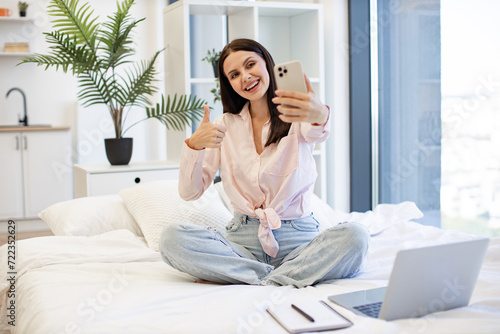 Caucasian young woman holding wireless smartphone having a video call showing thumb up. Beautiful lady sitting on comfy bed and enjoying online conversation with relatives or friends on weekend.