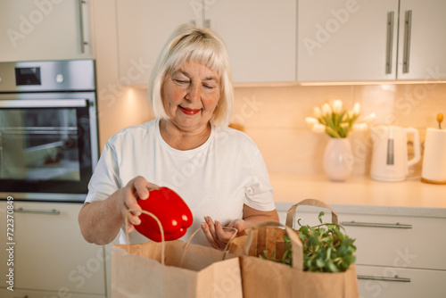 Happy lady unpacking groceries at home. Portrait of cheerful beautiful woman at kitchen table counter with bags full of apples, oranges, peppers, tomatoes, zucchinis, parsley from food market. High