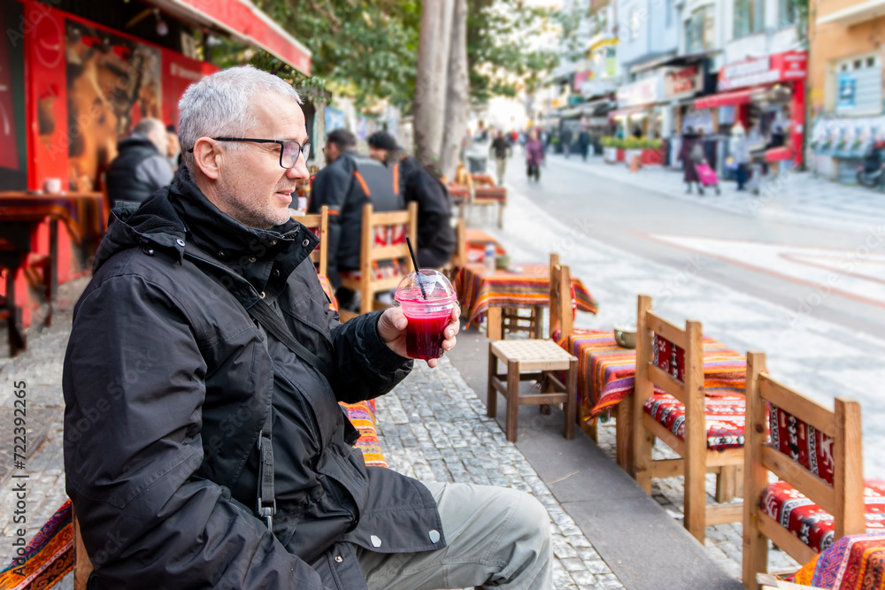 Obraz premium Male tourist drinking a pomegranate juice for refreshment. Portrait face of thirsty mature solo hipster traveler holding a pomegranate drink on street.