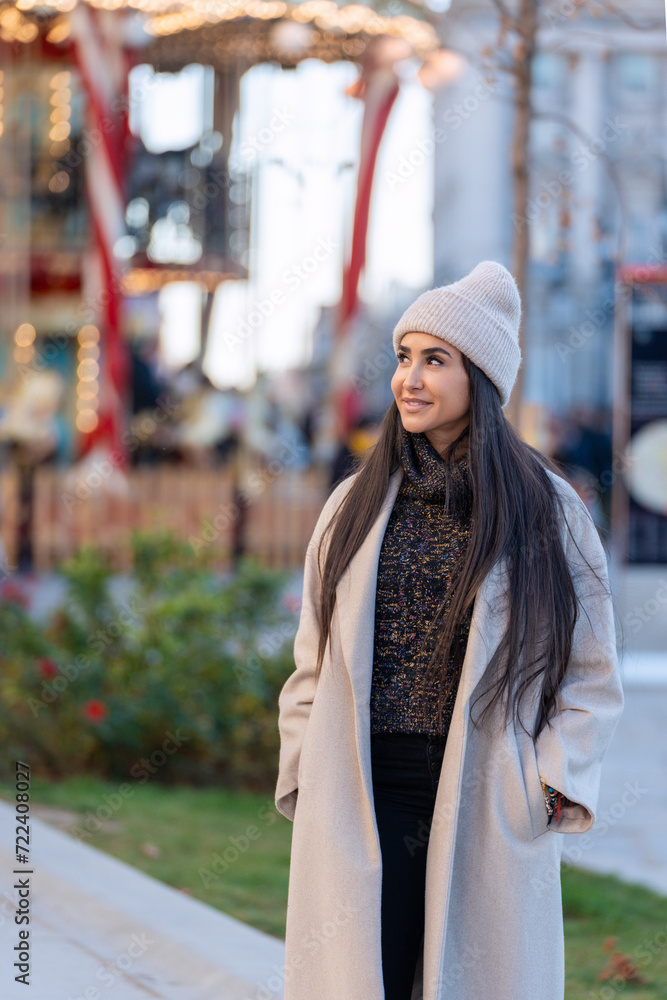 Smiling young woman in a stylish coat and beanie stands near a carousel in a festive winter setting, with soft focus lights in the background. Smiling young woman in a stylish coat and beanie stands near a carousel in a festive winter setting, with soft focus lights in the background.