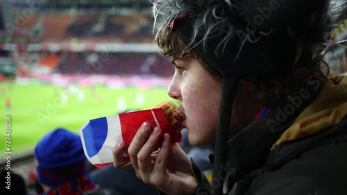 Teenager in a fur hat eating a hot dog at the stadium 