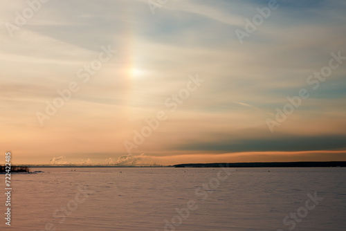 A winter rainbow (halo) over a frozen snow-covered river. In the distance a factory with many smoking pipes. Copy space.