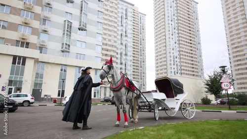 Coachman in black coat is holding horse by snaffle outside near skyscraper buildings photo