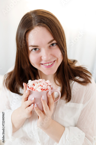 Beautiful woman with pink mug of cocoa and marshmallows on bed at home. St Valentine's Day.