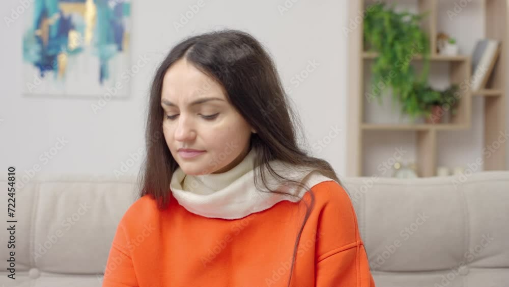 Young woman suffering from sore throat wearing scarf sitting on sofa sprays her throat suffering from pain Young woman suffering from sore throat wearing scarf sitting on sofa sprays her throat suffering from pain