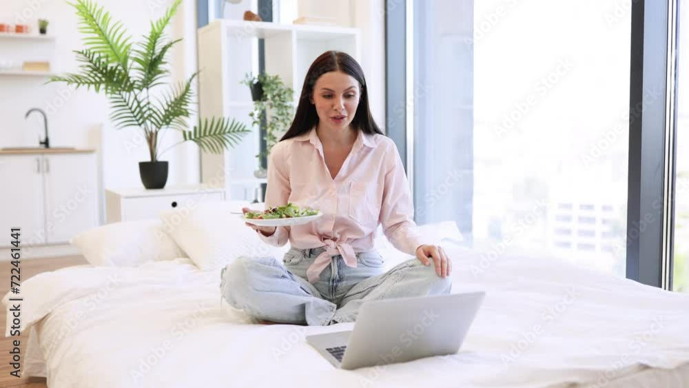 Front view of smiling woman having video chat with friends on laptop while eating healthy salad. Charming brunette resting on bed and enjoying nutrition from fresh vegetables while staying at home. Front view of smiling woman having video chat with friends on laptop while eating healthy salad. Charming brunette resting on bed and enjoying nutrition from fresh vegetables while staying at home.