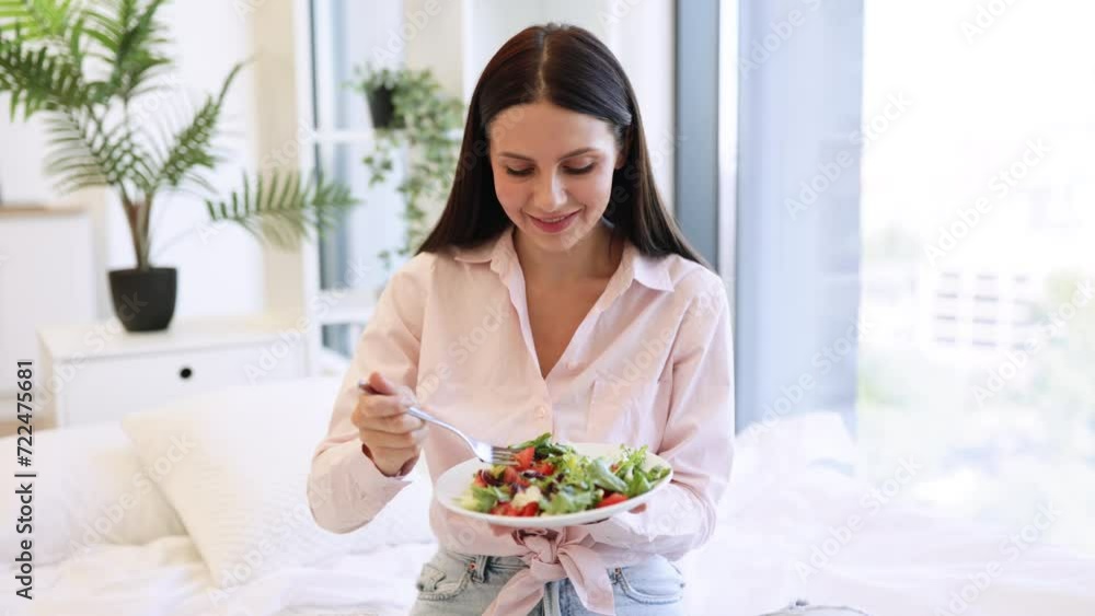 Close up portrait of caucasian woman in casual wear sitting on comfy bed eating healthy salad. Brunette female enjoying breakfast from fresh vegetables on background of light modern cozy bedroom. Close up portrait of caucasian woman in casual wear sitting on comfy bed eating healthy salad. Brunette female enjoying breakfast from fresh vegetables on background of light modern cozy bedroom.