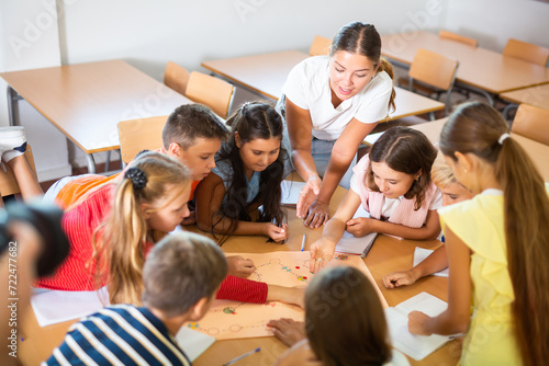 Happy preteen children and female teacher playing together educational board game in classroom at elementary school