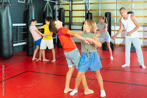 Kids exercising self-protection moves together on their training. Their teacher standing nearby and observing.