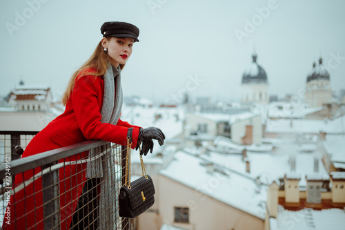 Fashionable woman wearing cap, red winter coat, gray scarf, gloves, holding black leather purse, posing on balcony with beautiful view on snow covered European city. Copy, empty, blank space for text