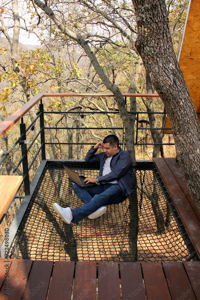 A 40-year-old dark-skinned Latin man uses his laptop on a net suspended over the forest on the terrace of his cottage, works as a digital nomad around the world