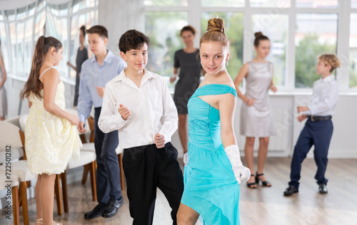 Smiling teenage girl in elegant dress having fun at school prom with group of peers, dancing with friend in spacious hall