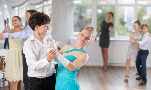 Happy interested teenage students preparing for college festive event, rehearsing ballroom dance steps in spacious classroom while female teacher observing from background..