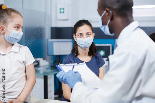 Caucasian woman and her daughter confer with male healthcare practitioner. African American physician seated on hospital bed  examines a little patient as everyone wears face masks.