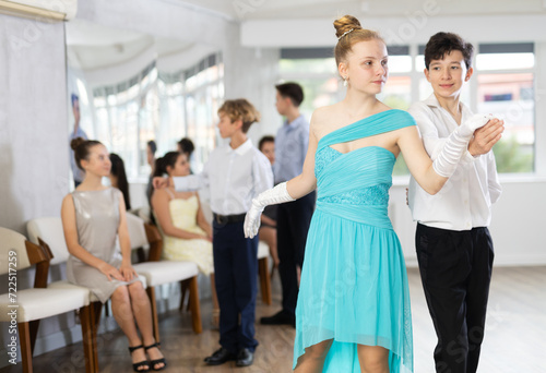 Enthusiastic friendly teenagers, boy and girl in party attire performing elegant waltz in pair in sunny hall of dance school during group lesson..