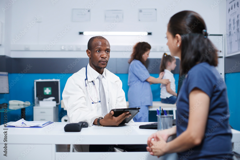 In hospital room, African American male doctor in a lab coat explains healthcare and medicine to a Caucasian woman. A female nurse performs a medical checkup on a small girl in the background. In hospital room, African American male doctor in a lab coat explains healthcare and medicine to a Caucasian woman. A female nurse performs a medical checkup on a small girl in the background.