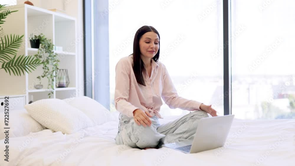 Happy brunette female waving hello while talking by video call using modern laptop, staying at home. Charming woman in casual attire enjoying online conversation with family while sitting on cozy bed. Happy brunette female waving hello while talking by video call using modern laptop, staying at home. Charming woman in casual attire enjoying online conversation with family while sitting on cozy bed.