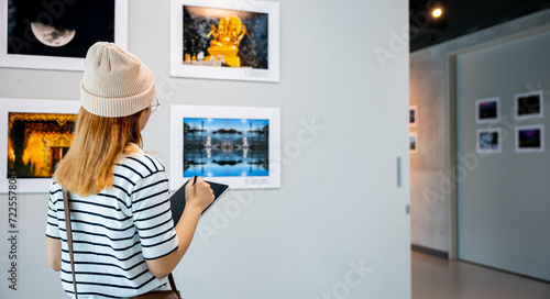 Young person at photo frame write digital book leaning against at show exhibit artwork gallery, Asian woman holding tablet at art gallery collection in front framed paintings looking pictures on wall