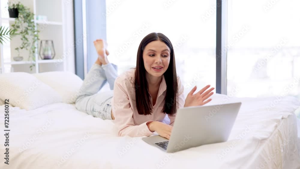 Happy brunette female waving hello while talking by video call using modern laptop, staying at home. Charming woman in casual attire enjoying online conversation with family while lying on cozy bed. Happy brunette female waving hello while talking by video call using modern laptop, staying at home. Charming woman in casual attire enjoying online conversation with family while lying on cozy bed.