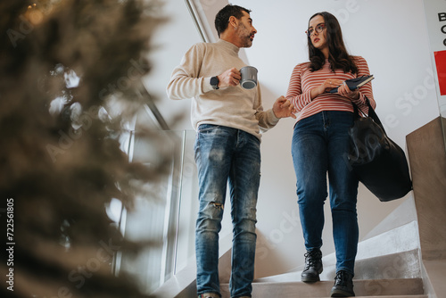 Billede på lærred Two creative colleagues engaging in a casual chat while holding coffee, standing beside a staircase with a subtle Christmas tree blur