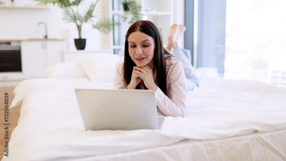 Charming woman in casual attire enjoying online conversation with friends while lying on cozy bed. Happy brunette female gesturing while talking by video call using modern laptop, staying at home. Charming woman in casual attire enjoying online conversation with friends while lying on cozy bed. Happy brunette female gesturing while talking by video call using modern laptop, staying at home.