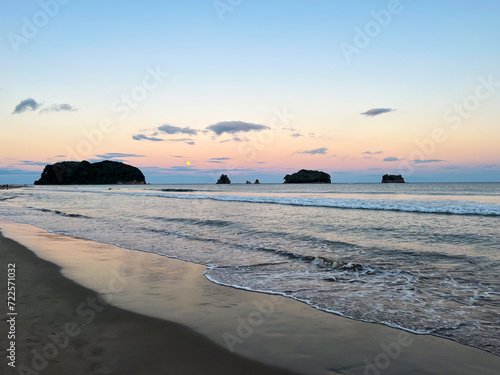 Whangamata Beach, where the sun gracefully sets, casting warm hues across the sky, and a full moon rises