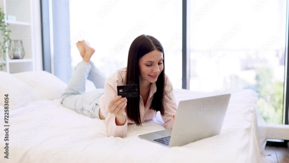 Smiling young woman making online order using modern wireless laptop. Relaxed lady lying on comfy bed and enjoying purchase of goods on Internet using bank card at home on weekend. Smiling young woman making online order using modern wireless laptop. Relaxed lady lying on comfy bed and enjoying purchase of goods on Internet using bank card at home on weekend.