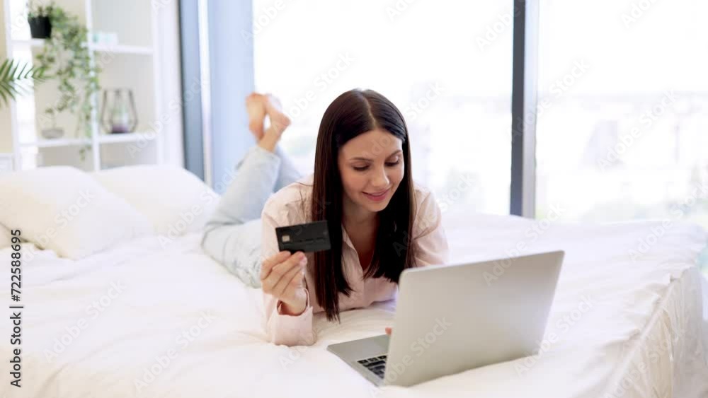Brunette female typing on laptop entering credit card information for purchase, staying at home looking at camera. Charming woman in casual attire enjoying online shopping while lying on cozy bed. Brunette female typing on laptop entering credit card information for purchase, staying at home looking at camera. Charming woman in casual attire enjoying online shopping while lying on cozy bed.