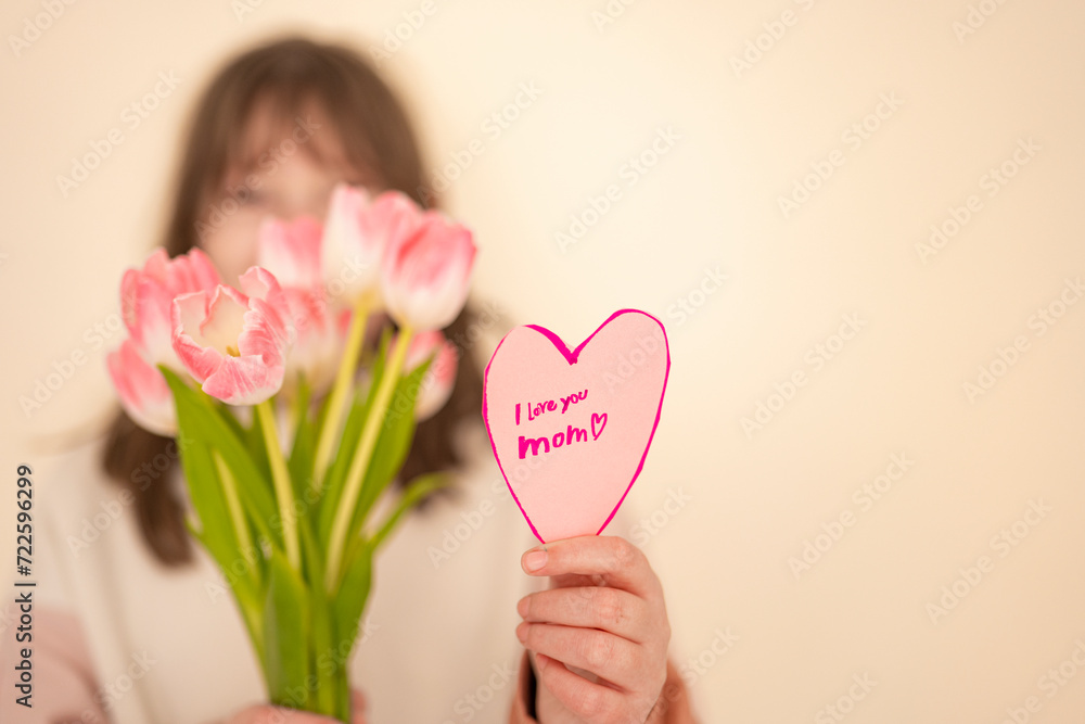 Mothers Day.daughter with flowers and card on a beige background.Laughing girl with tulips and heart card.Pink heart card and pink tulips in the hands of a girl.moms day.Message to mom. Mothers Day.daughter with flowers and card on a beige background.Laughing girl with tulips and heart card.Pink heart card and pink tulips in the hands of a girl.moms day.Message to mom.