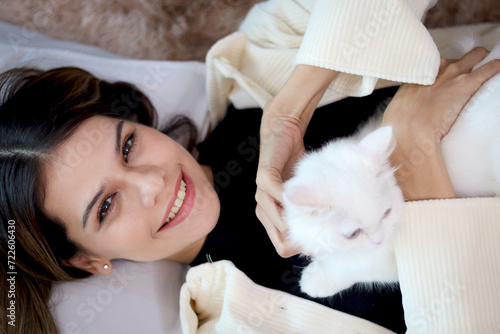 Fototapeta Naklejka Na Ścianę i Meble -  Portrait of young happy smiling woman comfortably lying on sofa in living room with cute white Persian cat sitting on her, girl gentle playing with fluffy long hair kitty pet with love.