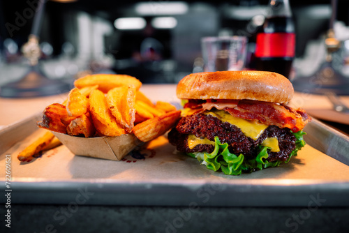 A delicious hamburger with two beef patties, bacon, lettuce, cheese and brioche bun, accompanied by chunky waffle cut fries served on a tray with restaurant kitchen, glass & drink in bokeh background photo