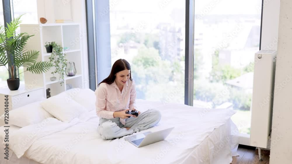 Happy lady sitting on comfy bed and celebrating victory in favorite computer game on weekend. Young woman raising hands with wireless joystick while win in video games using modern laptop. Happy lady sitting on comfy bed and celebrating victory in favorite computer game on weekend. Young woman raising hands with wireless joystick while win in video games using modern laptop.