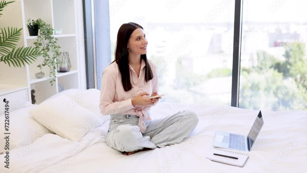 Relaxed lady sitting on comfy bed and enjoying chatting with coworkers using smartphone on weekend. Attractive young woman working remotely from home, typing on modern wireless cell phone. Relaxed lady sitting on comfy bed and enjoying chatting with coworkers using smartphone on weekend. Attractive young woman working remotely from home, typing on modern wireless cell phone.