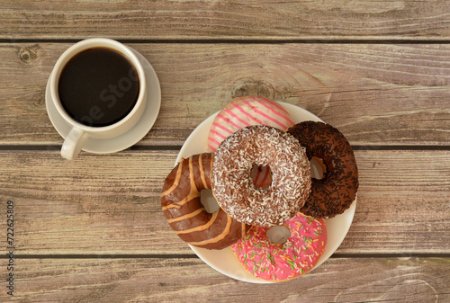 A cup of hot black coffee on a saucer and a plate with a bunch of fresh donuts in multi-colored glaze on a wooden table.