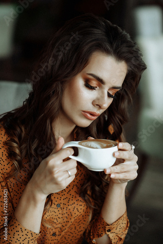 A serene Caucasian woman with wavy hair, in a polka-dotted blouse, savoring her coffee in a cozy, softly lit interior. Front view. Concept for a tranquil morning routine.