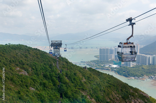 View of gondolas of Ngong Ping Cable Car (Skyrail) gliding over the seaside hills with the airport on Chek Lap Kok island & residential towers on the hillside under sunny cloudy sky in Hong Kong China
