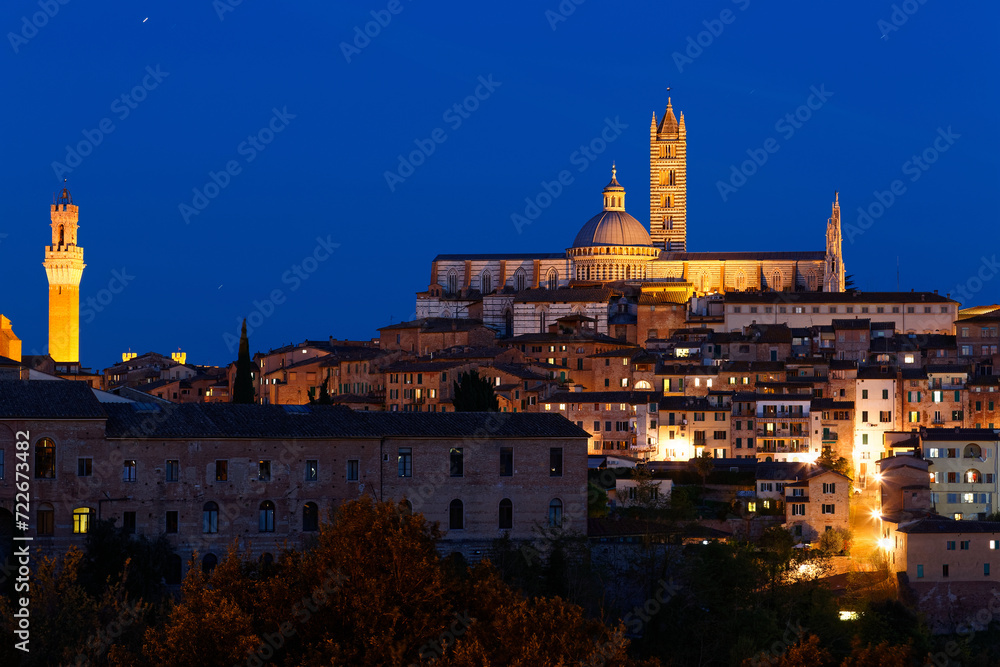 Night scene of Siena, a medieval town on a hilltop in Tuscany, Italy, with a view of architectural landmarks (Dome & Bell Tower of Siena Cathedral and Mangia Tower) lighted up in blue evening twilight